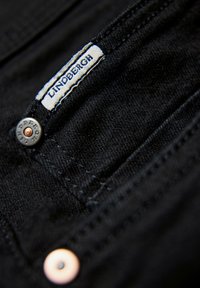 Gros plan sur un tissu en denim noir avec une étiquette de la marque LINDBERGH et deux rivets métalliques sur le coin de la poche.