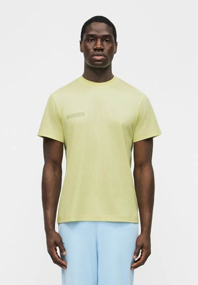 Άνδρας που στέκεται με το πρόσωπο προς τα εμπρός, φορώντας ανοιχτό κίτρινο κοντομάνικο μπλουζάκι με μικρό κείμενο στο στήθος και ανοιχτό μπλε παντελόνι.