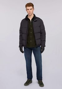 Svart vadderad jacka med horisontellt quiltat mönster, dragkedja framtill och tryckknapp vid kragen. Bärs över en mörkgrön skjorta, kombinerad med blå jeans och gröna sneakers.