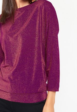 Femme portant un haut à manches longues violet scintillant, coupé à la taille, avec un pantalon noir visible sur un fond blanc uni.