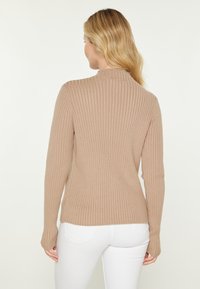 Rippiger beige Pullover mit hohem Kragen, langen Ärmeln und enger Silhouette. Weicher Stoff mit leichtem Stretch, getragen zu weißen Jeans.