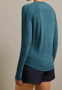 Langärmliges sportliches Shirt in Teal mit strukturiertem Stoff, Raglanärmeln und Flachnähte. Ansicht von hinten, kombiniert mit dunklen Shorts.