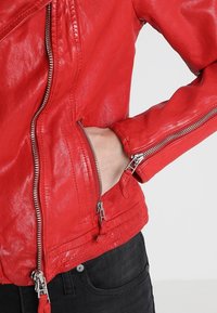 Hand in zijzak van rode leren jas met zilveren ritsdetails, gedragen over zwarte broek tegen een lichte achtergrond.