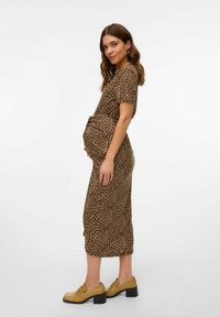 Vestido de leopardo con mangas cortas y cinturón en la cintura, de tejido fluido y con fruncidos laterales para una forma ajustada sobre una silueta de embarazo. Llevado con zapatos amarillos de tacón cuadrado.