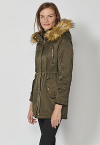 Olivovo zelený parka kabát s kapucňou lemovanou kožušinou, s vreckami na zips a gombíky, elastickým pásom a kontrastným čiernym lemovaním. Hladká textúra tkaniny.