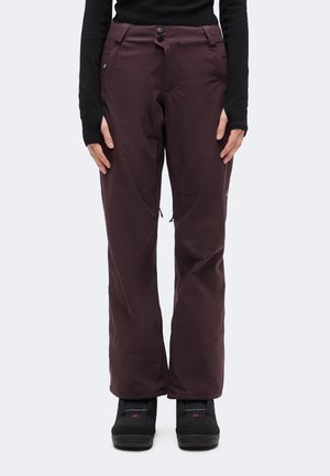 Personne portant un pantalon de neige violet foncé et des bottes noires, debout les mains détendues le long du corps sur un fond clair uni.