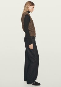 Braune Strickweste über einem schwarzen Rollkragenpullover, kombiniert mit dunkelblauen Jeans mit weitem Bein und dunklen Schuhen. Seitenansicht, Haare offen.