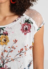 Bluzka krótki rękaw w jasnoszarym kolorze z kwiatowym wzorem. Posiada siateczkowe ramiączka w kropki oraz koronkowe wykończenia. Oddychający materiał.