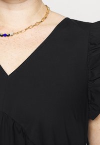 Gros plan sur une personne portant une robe noire à col en V avec des manches à volants et un collier en chaîne dorée orné de perles bleues et noires.