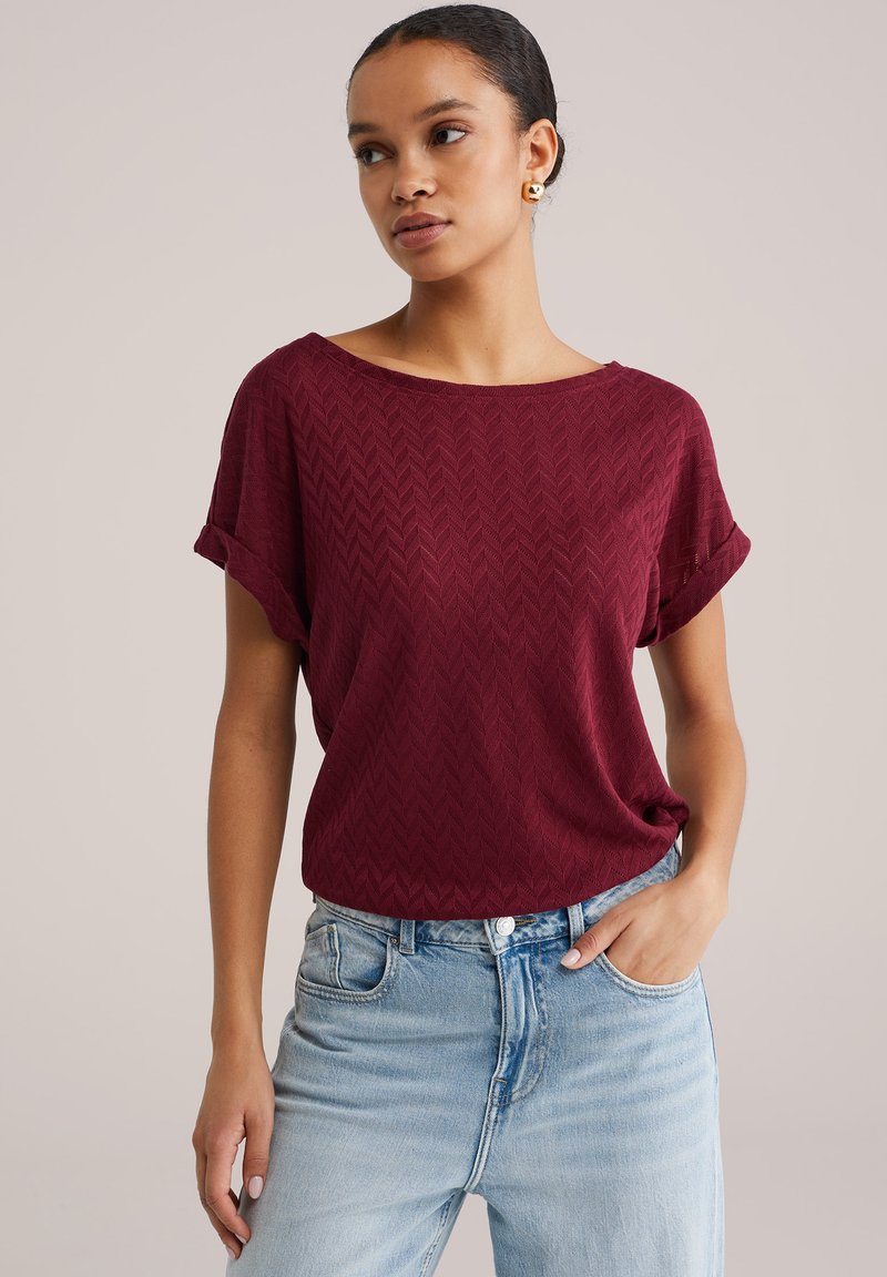 Burgundska majica kratkih rokavov z teksturiranim vzorcem ribje kosti, sproščenim oprijetjem in zavihanimi rokavi, v kombinaciji s svetlo modrimi visokimi kavbojkami.