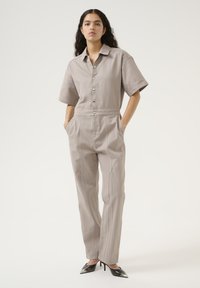 Beige kortärmad jumpsuit med knäppning fram, elastisk midja, sidofickor och dekorativa nitar på axlarna. Kombineras med svarta klackskor.