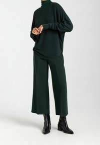 Ciemnozielony, oversize'owy sweter dzianinowy z wysokim kołnierzem i obniżonymi ramionami, zestawiony z szerszymi spodniami w pasującym materiale, noszonymi z czarnymi botkami za kostkę.
