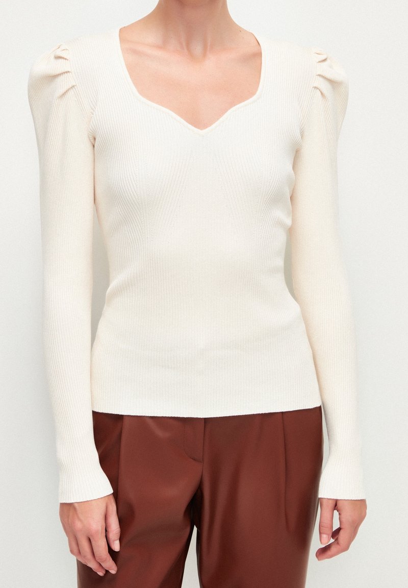 Femme portant un pull crème côtelé à manches longues avec des épaules bouffantes et un pantalon plissé marron sur un fond blanc uni.