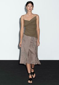 Olivengrønt fitted top med draperingsdetalje, parret med en taupe ruskind indpakningsskørt. Sorte flade sandaler med flere stropper. Minimalistisk baggrund.