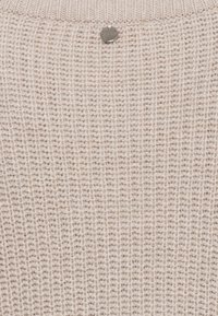 Maglione lavorato a maglia beige con un motivo strutturato, scollo tondo e un piccolo logo metallico vicino al colletto.