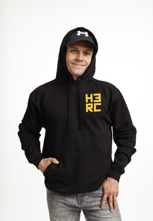 Mężczyzna w czarnej bluzie z kapturem z żółtym logo "H3RC" i czarnej czapce, uśmiechający się z jedną ręką w kieszeni, na jednolitym białym tle.