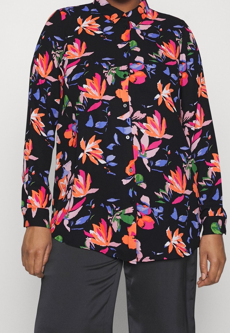 Chemise noire à manches longues avec un motif floral vif en orange, rose, violet et vert, portée avec un pantalon sombre.