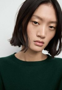 Primo piano di una persona con capelli scuri di lunghezza spalle, che indossa un maglione verde con scollo rotondo, che mostra una pelle liscia e un'espressione neutra.