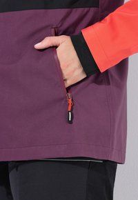Giacca viola con accenti neri, con una mano in una tasca laterale. La cerniera della tasca è dotata di un tirazip arancione per un facile accesso.