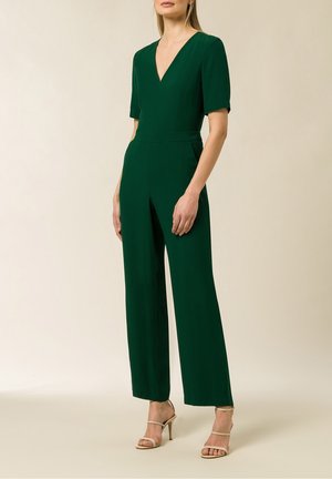 Mujer con un mono verde oscuro de manga corta y escote en V profundo, combinado con sandalias de tacón alto beige, de pie frente a un fondo neutro.