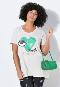 Camiseta beige claro con un gráfico de un corazón y un ojo verdes, con la palabra "Happy" en dorado. Combinada con un bolso tejido verde y unos vaqueros negros desgastados.