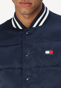 Chaqueta acolchada navy con cuello acanalado de rayas blancas, botones a presión y un pequeño logo bordado de "TOMMY JEANS" en el pecho.