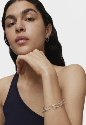 Pulsera de plata con formas circulares enlazadas; la modelo lleva una camiseta sin mangas azul marino, mostrando la pulsera en un brazo al descubierto. Textura de piel suave.