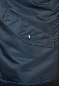 Poche en tissu bleu foncé avec un rabat triangulaire fixé par un seul bouton pression rond sur la manche d'une veste.