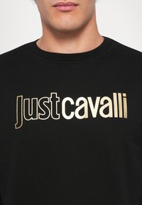 Muž v čiernom svetri s okrúhlym výstrihom a veľkým zlatým logom "Just Cavalli" na hrudi, zobrazený od pier po hornú časť trupu.