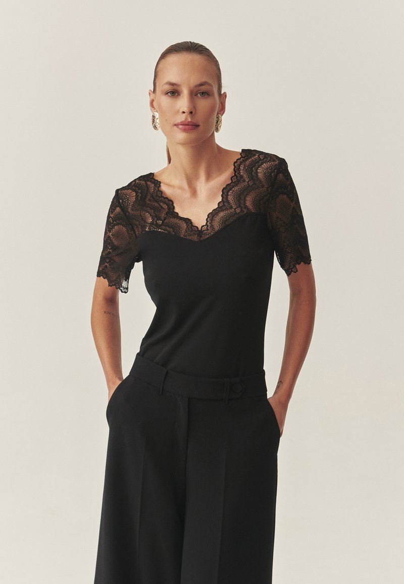 Črna majica s kratkimi rokavi, ki ima čipkasto oblogo na izrezu in rokavih. Ujemajoče se s krojenimi črnimi hlačami. Gladek tekstura tkanine.