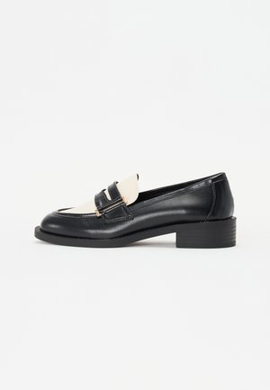 Black leather loafer with a contrasting cream vamp, featuring two decorative straps with gold hardware and a block heel design.