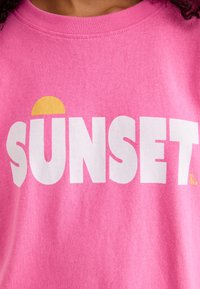 Růžové bavlněné tričko s výraznými bílými písmeny, které hlásí "SUNSET" a žlutým poloobvodem nad textem.