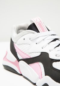 Close-up of white, pink, and black Puma sneaker with textured fabric, thick sole, white laces, and pink Puma logo on tongue.