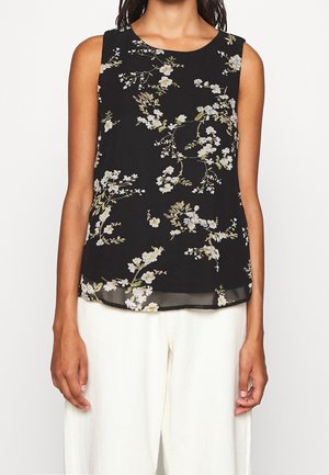 Femme portant un haut noir sans manches avec un motif floral blanc et jaune, assorti à un pantalon blanc ample, sur un fond uni.