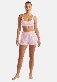 Kobieta stojąca boso, w jasnoróżowym staniku sportowym i pasujących szortach na tle białym.