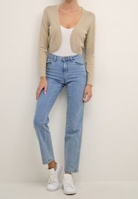 Beżowy skrócony kardigan na biały top, w połączeniu z jasnoniebieskimi spodniami o wysokim stanie i białymi sneakersami. Gładka tekstura, swobodny krój.