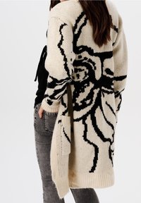 Beige pletený kardigan s černým květinovým vzorem. Obsahuje otevřenou přední část, dvě boční kapsy a žebrované manžety a lem. Neformální design.
