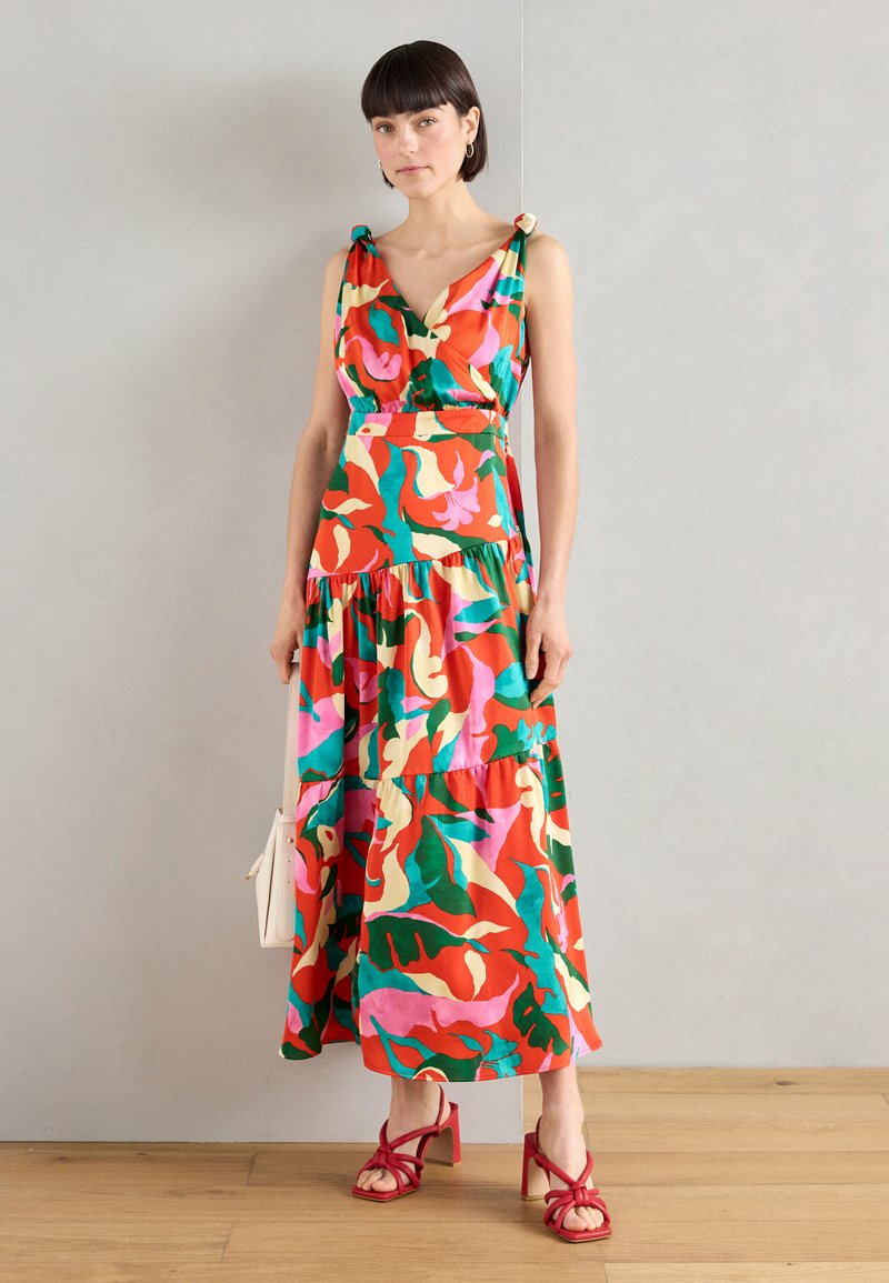 Robe maxi florale avec des motifs vibrants rouges, verts et roses, dotée d'un décolleté en V et d'une jupe à volants. Associée à des sandales rouges et un sac blanc.