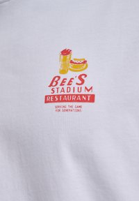 Logo z napojem i kanapką nad tekstem „Bee's Stadium Restaurant” oraz hasłem „Serving the game for generations” na białej tkaninie.