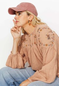 Casquette en velours côtelé rose, blouse beige brodée avec des manches bouffantes, et jeans délavés bleu clair. Boucles d'oreilles en or visibles.