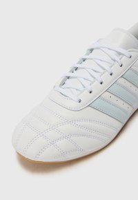 Hvide lædersoccer sko med lyseblå detaljer, der har en tekstureret overdel, kontrasterende syninger og gummisål. Snørebånd og ventilationshuller inkluderet.
