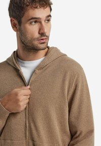 Mann mit kurzem braunem Haar, der einen beige farbigen Reißverschluss-Hoodie über einem weißen Shirt trägt, zur Seite schaut und den Reißverschluss hält.