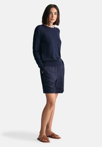 Suéter de punto azul marino con puños acanalados, combinado con pantalones cortos de lino azul marino con bolsillos laterales. Sandalias planas marrones con acentos decorativos.