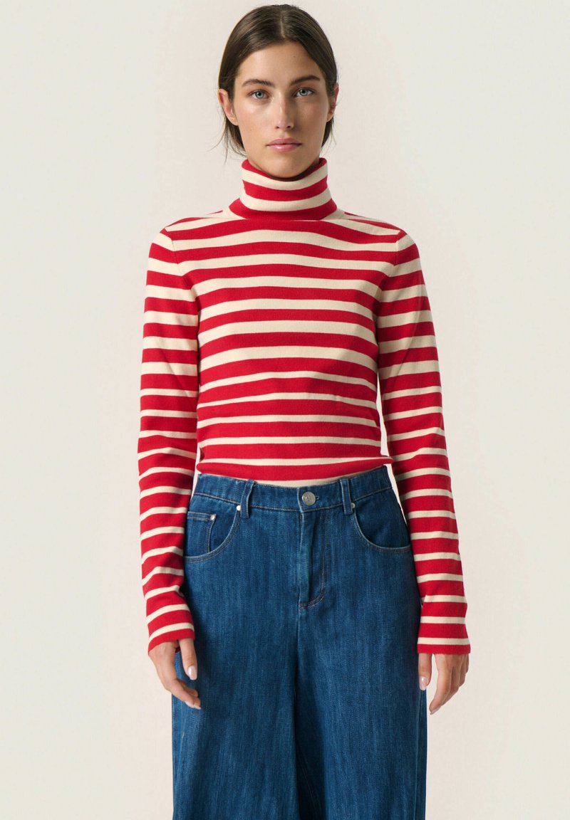 Maglione a collo alto a righe rosse e crema abbinato a pantaloni in denim blu a vita alta e a gamba larga. Texture a maglia liscia e maniche aderenti.