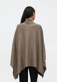 Γυναίκ�α με μακριά σπαστά μαλλιά, ντυμένη με καμηλό τζάκετ με γιακά και μαύρο παντελόνι, φαίνεται από πίσω σε απλό φόντο.