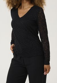 Top lungo manica in pizzo nero con scollatura a V, caratterizzato da intricate fantasie floreali e maniche trasparenti, abbinato a pantaloni neri.