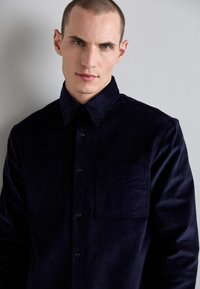 Camisa de pana en color azul marino con cuello puntiagudo, cierre frontal de botones y dos bolsillos en el pecho, que presenta un acabado suave y texturizado.