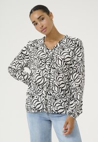Black and white patterned blouse with a V-neck, ruffled front, long sleeves, and soft, textured fabric. Paired with denim jeans.