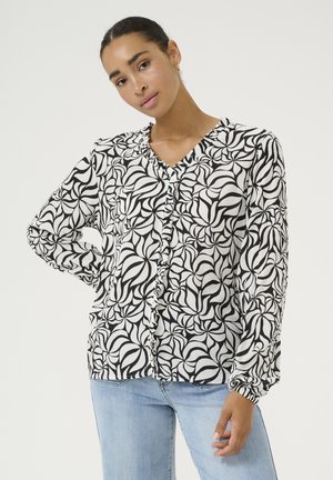 Blouse à motifs noir et blanc avec un décolleté en V, un devant froncé, des manches longues et un tissu doux et texturé. Associée à un jean en denim.
