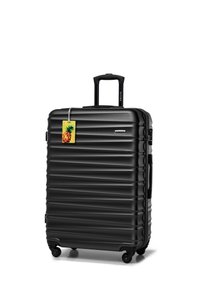 Valise rigide noire avec des rainures verticales, poignée rétractable et quatre roues pivotantes. Un tag à bagages en forme d'ananas coloré est attaché.